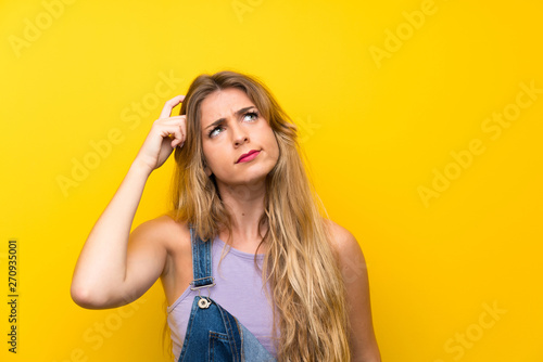 Young blonde woman with overalls over isolated yellow background having doubts and with confuse face expression