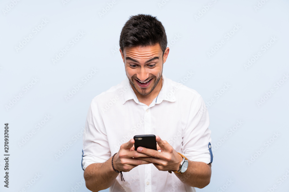 Handsome young man over isolated blue background surprised and sending a message Handsome young man over isolated blue background surprised and sending a message