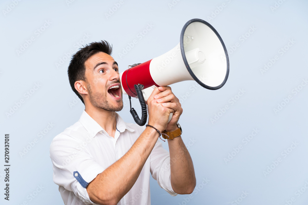 Handsome young man over isolated blue background shouting through a megaphone Handsome young man over isolated blue background shouting through a megaphone