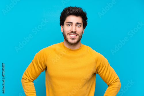 Handsome over isolated blue wall posing with arms at hip and smiling