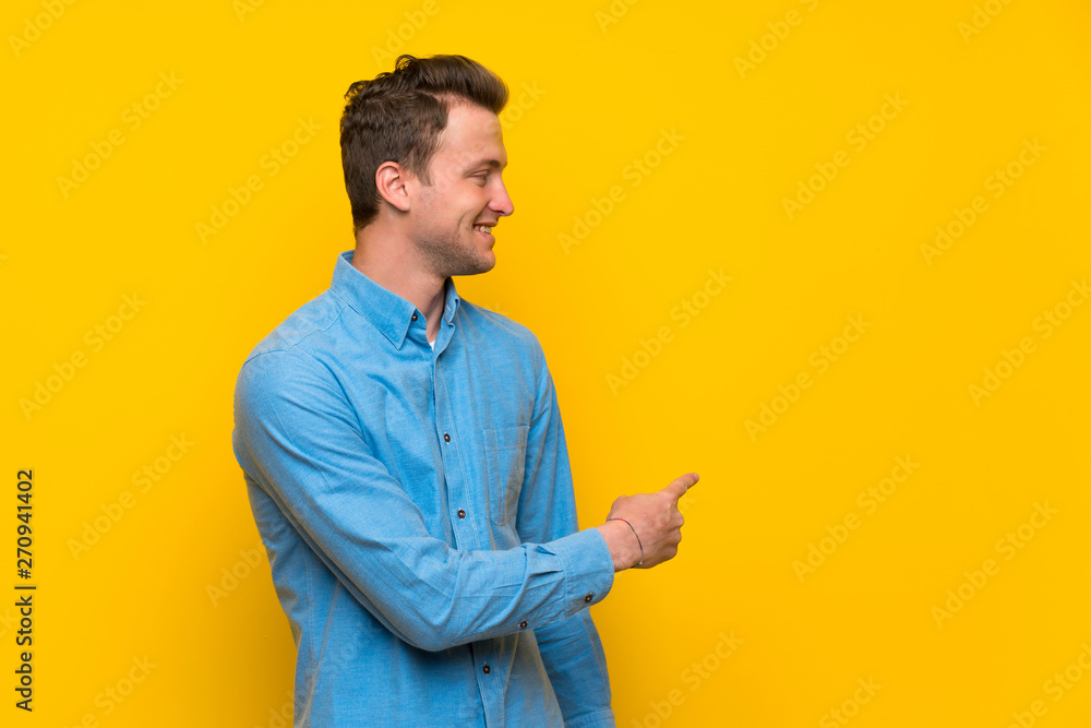 Blonde man over isolated yellow wall pointing back Blonde man over isolated yellow wall pointing back