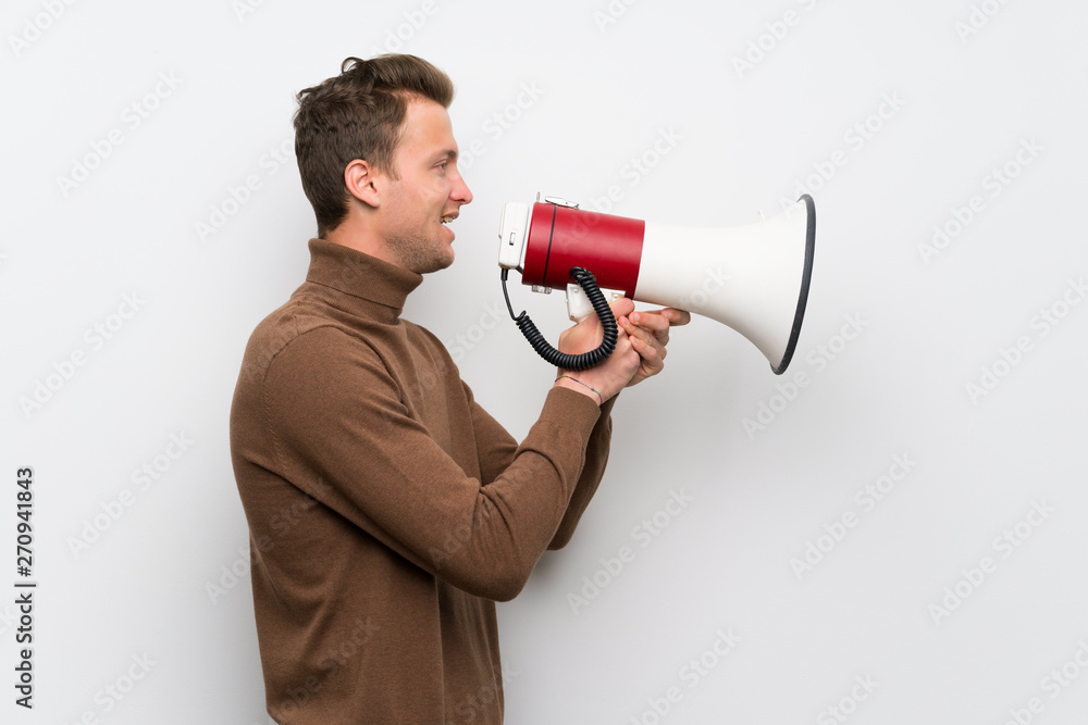 Blonde man over isolated white wall shouting through a megaphone Blonde man over isolated white wall shouting through a megaphone