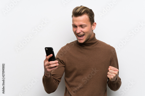Blonde man over isolated white wall celebrating a victory with a mobile © luismolinero