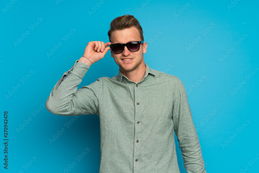 Blonde man over isolated blue wall with glasses and surprised Blonde man over isolated blue wall with glasses and surprised