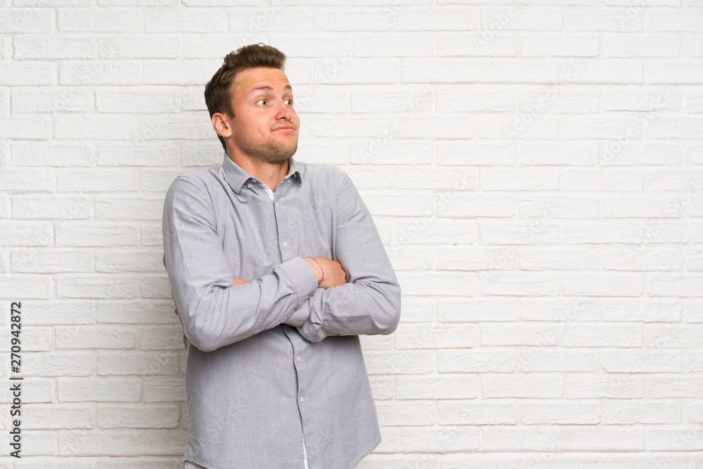 Blonde man over white brick wall making doubts gesture while lifting the shoulders Blonde man over white brick wall making doubts gesture while lifting the shoulders