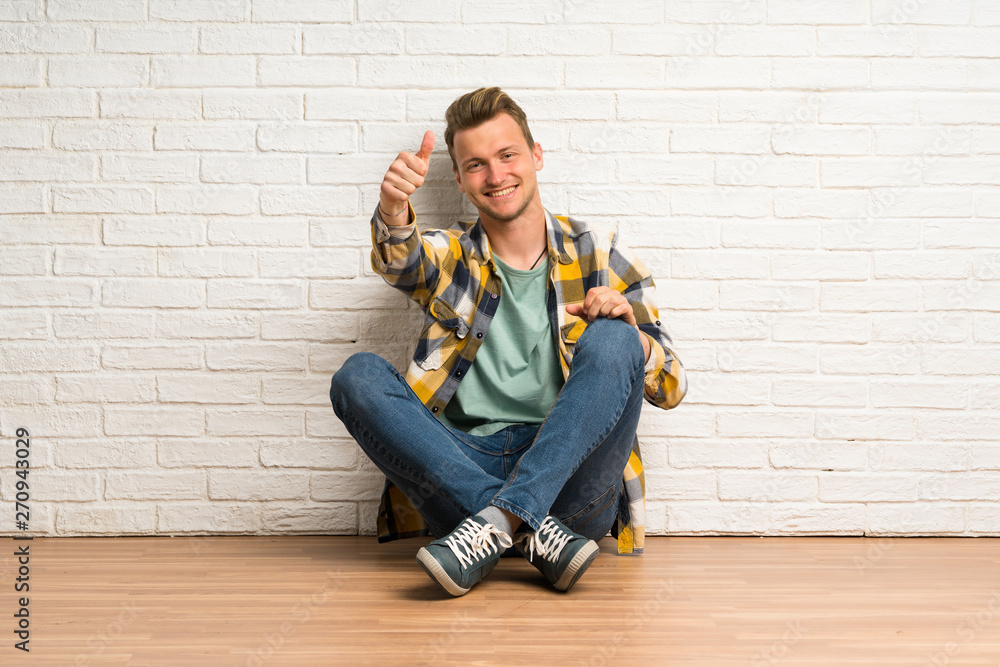 Blonde man sitting on the floor with thumbs up because something good has happened Blonde man sitting on the floor with thumbs up because something good has happened