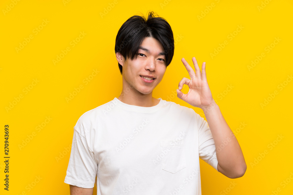 Asian man over isolated yellow wall showing ok sign with fingers Asian man over isolated yellow wall showing ok sign with fingers