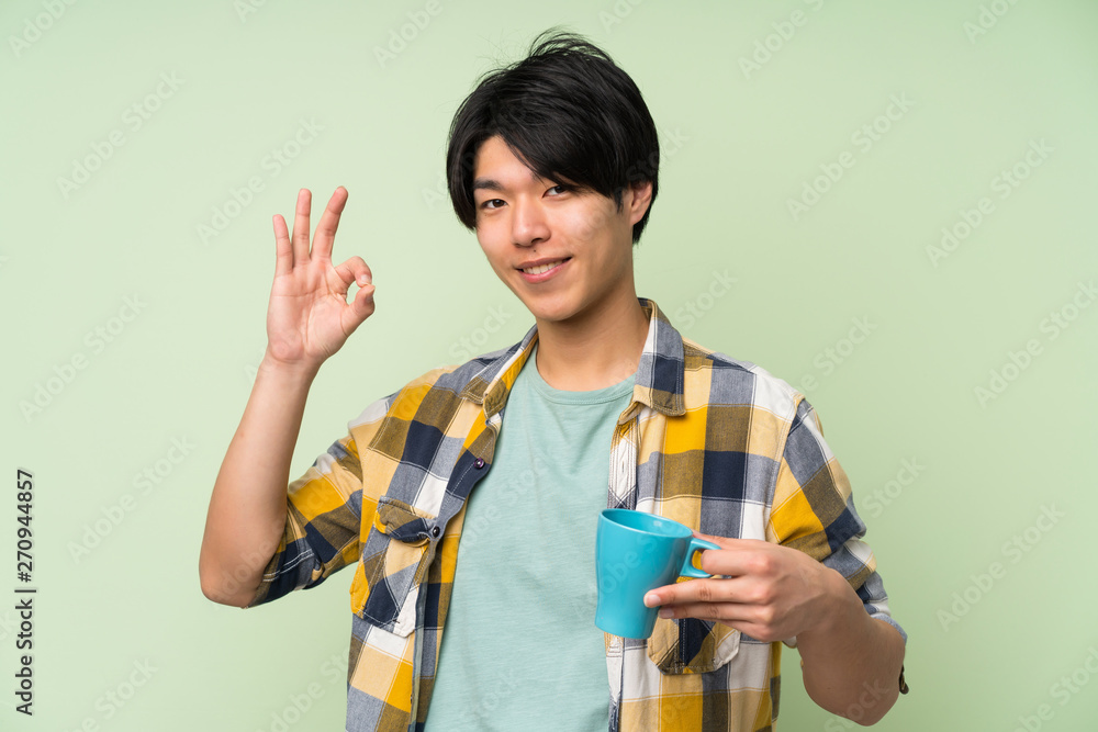 Asian man over isolated green wall holding hot cup of coffee Asian man over isolated green wall holding hot cup of coffee