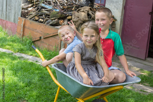 Vászonkép kids in wheelbarrow,two boys and a girl were sitting in a wheelbarrow in the sum