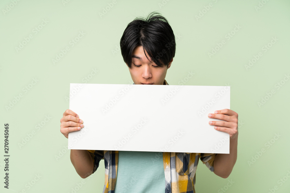 Asian man over isolated green wall holding an empty white placard for insert a concept Asian man over isolated green wall holding an empty white placard for insert a concept