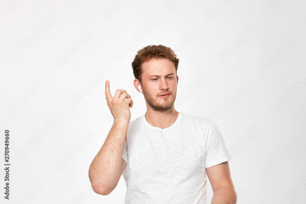 Portrait of a bearded handsome young man with wireless headphones holding hand near headset trying to hear sound standing infront of white bakground. Portrait of a bearded handsome young man with wireless headphones holding hand near headset trying to hear sound standing infront of white bakground.