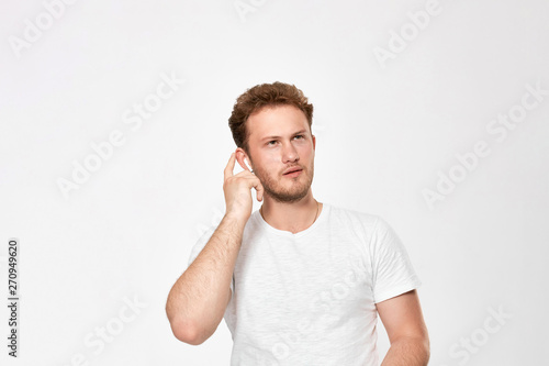 Portrait of a bearded handsome young man with wireless headphones holding hand near headset trying to hear sound standing infront of white bakground.