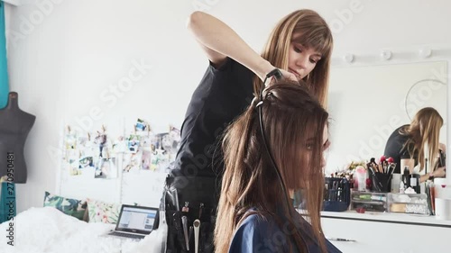 Timelapse shot of cute young female barber enjoying working with a beautiful young brunette model, doing fresh trendy hairstyle on her long curly hair using curling iron, hairbrush, hair spray