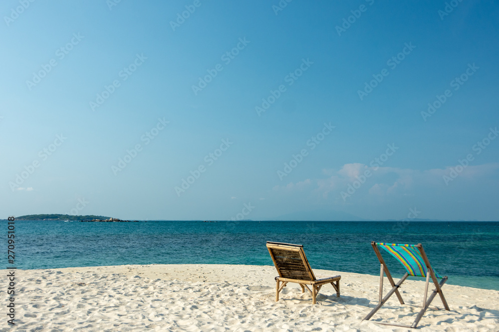 Deckchair and Wooden chairs on the white sandy beach with little waves, blue and bright blue sky.