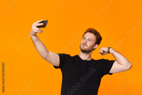 Studio shot of a smiling bearded young man making selfie with the smartphone while standing over orange background