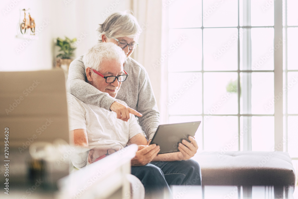 couple of seniors smiling and looking at the tablet - woman hogging at man with love on the sofa - indoor - showing couple of seniors smiling and looking at the tablet - woman hogging at man with love on the sofa - indoor - showing