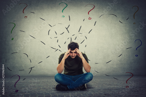 Photos Confused and tired young man seated on the floor keeps hands to forehead looking down thoughtful surrounded by different tasks to solve as having many questions