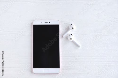 Smartphone with white wireless earphones with selective focus on white wooden background with empty space for text. Mobile phone and portable headphones on textured backdrop 
