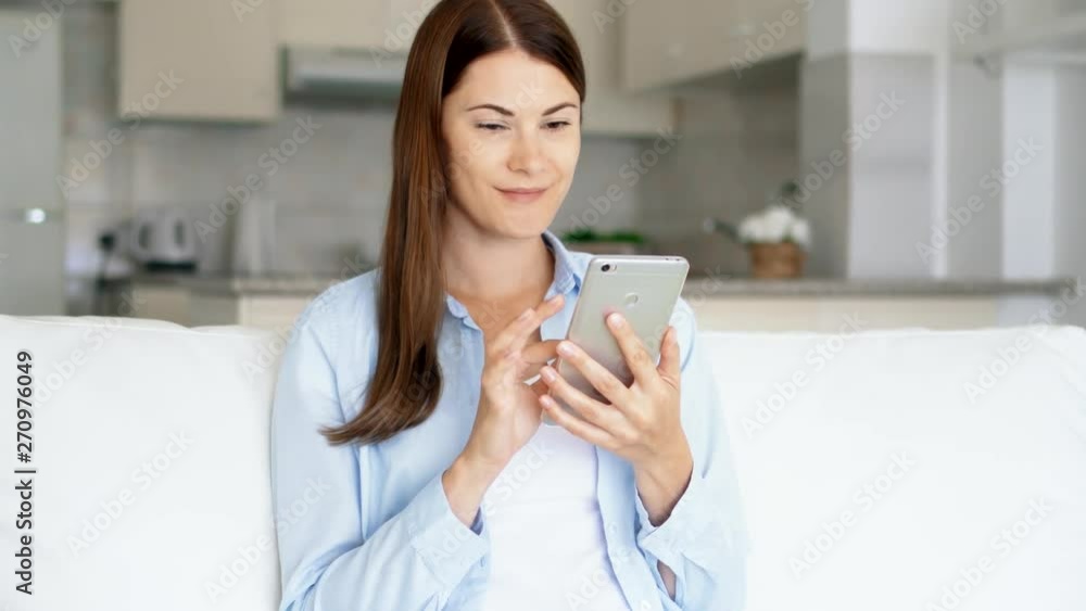 Young brunette woman with brown eyes sitting on sofa in living room using smartphone. Female housewife browsing on mobile phone chatting online with friends. Kitchen on background Young brunette woman with brown eyes sitting on sofa in living room using smartphone. Female housewife browsing on mobile phone chatting online with friends. Kitchen on background