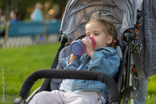 Little Caucasian girl dressed in a denim jacket drinks juice from her bottle while sitting in a baby carriage