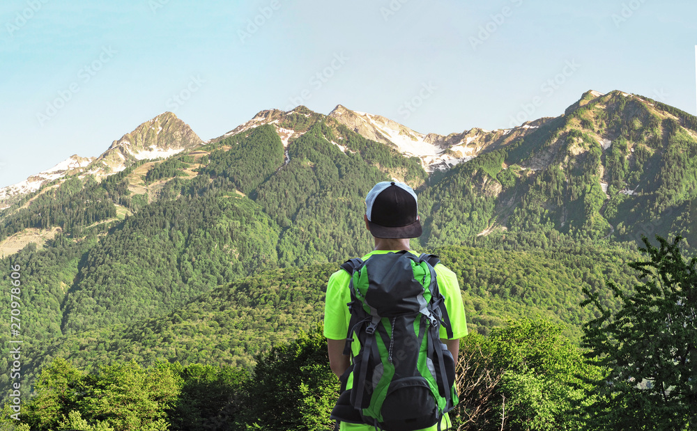Back view of young active man standing at mountain range background at sunny summer day. Person with backpack on trekking