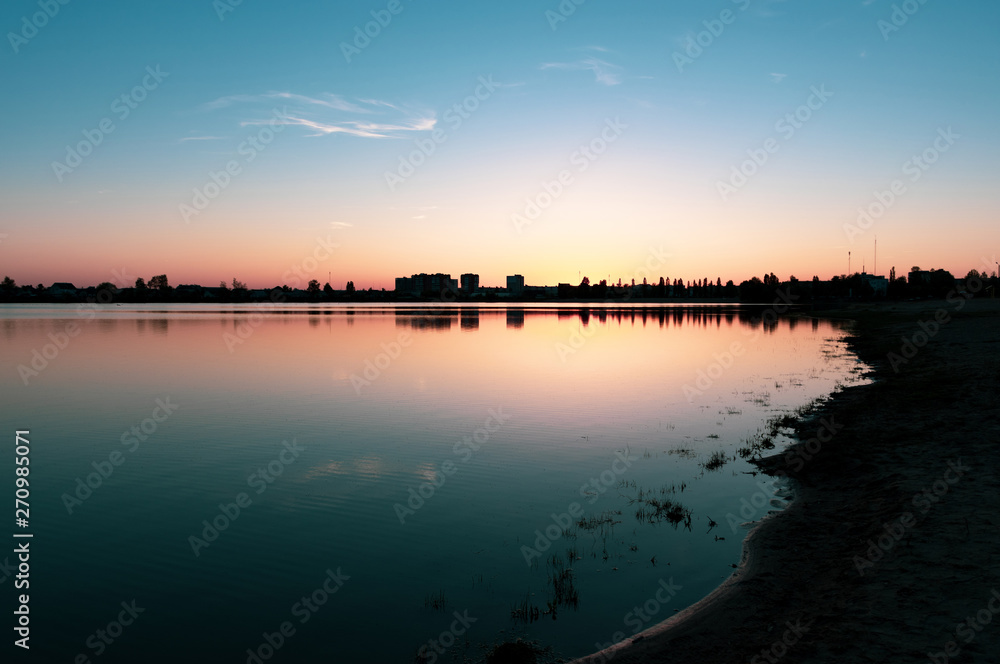 Sunset above the lake and city beach in summer with dark silhouettes of houses