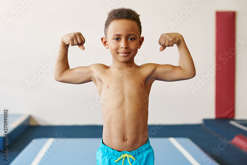 Children, fitness, health and ethnicity concept. Portrait of dark skinned black boy of school age exercising at gym, preparing for competition, posing topless, demonstrating his tensed muscles photo