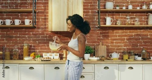 Cheerful young African American charming girl in pajama shaking eggs in a bowl while cooking in the kitchen, dancing and having fun. photo
