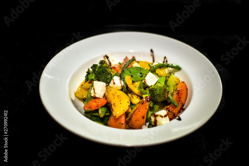 a white plate setup showing a delicious home made salad consisting of a variety of vegetables like capsicums, cottage cheese, broccoli, tomatoes, baby corns etc served with a sauce dressing