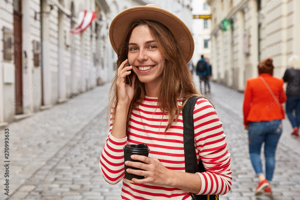 Happy traveler calls operator via cellphone, listens internet benefits, stands outdoor against blurred street background, wears stylish hat, has trip in urban setting, looks gladfully at camera