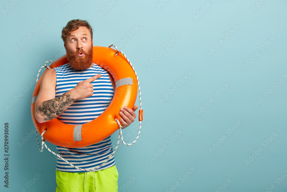 Shot of puzzled surprised redhaired male rescuer with lifebuoy, shows direction, points at upper right corner, tells impressive story about saving sinking person. Man on awesome summer vacation - obrazy, fototapety, plakaty 