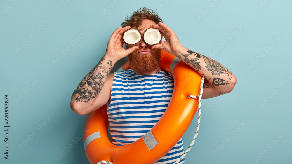 Curly red haired man keeps cocnonut on eyes, has fun near water, feels bored working as liveguard, wears sailor vest, stands over blue background, carries orange lifebuoy for saving people at beach - obrazy, fototapety, plakaty 