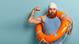 Self confident strong male lifeguard shows muscles, raises arm, has serious expression, carries safety ring, cares about water safety, helps swimmers who are in trouble, responds on aquatic emergency