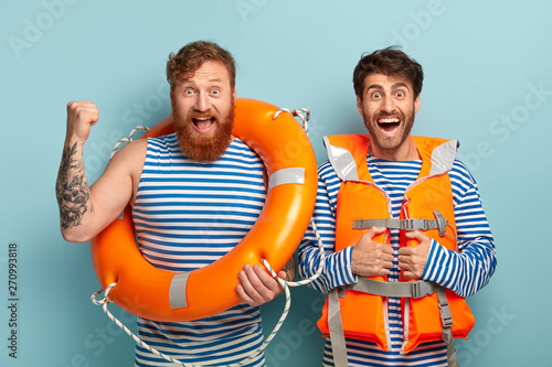 Two men lifeguards use lifeline, wear special orange vest, look happily at camera, saved life of swimmer at beach, does their duties, keep beach safe in summertime period. Life savers watch sea