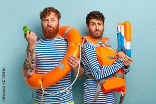 Serious rescue team wears life vest, hold lifebuoy or lifepreserver, waterguns, ready to save swimmers, watch sea attentively, care about salvation, stand shoulder to shoulder against blue background. photo