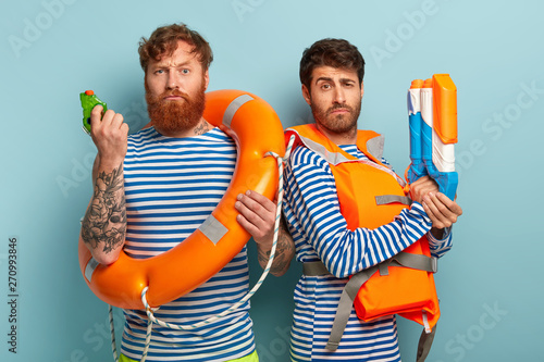 Serious rescue team wears life vest, hold lifebuoy or lifepreserver, waterguns, ready to save swimmers, watch sea attentively, care about salvation, stand shoulder to shoulder against blue background.