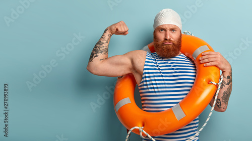 Self confident strong male lifeguard shows muscles, raises arm, has serious expression, carries safety ring, cares about water safety, helps swimmers who are in trouble, responds on aquatic emergency