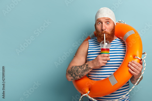 Embarrassed holiday maker drinks cold summer cocktail, spends free time at beach, wears swimcap sailor t shirt, swims with lifebuoy, has surprised expression, models over blue wall with free space #270993899