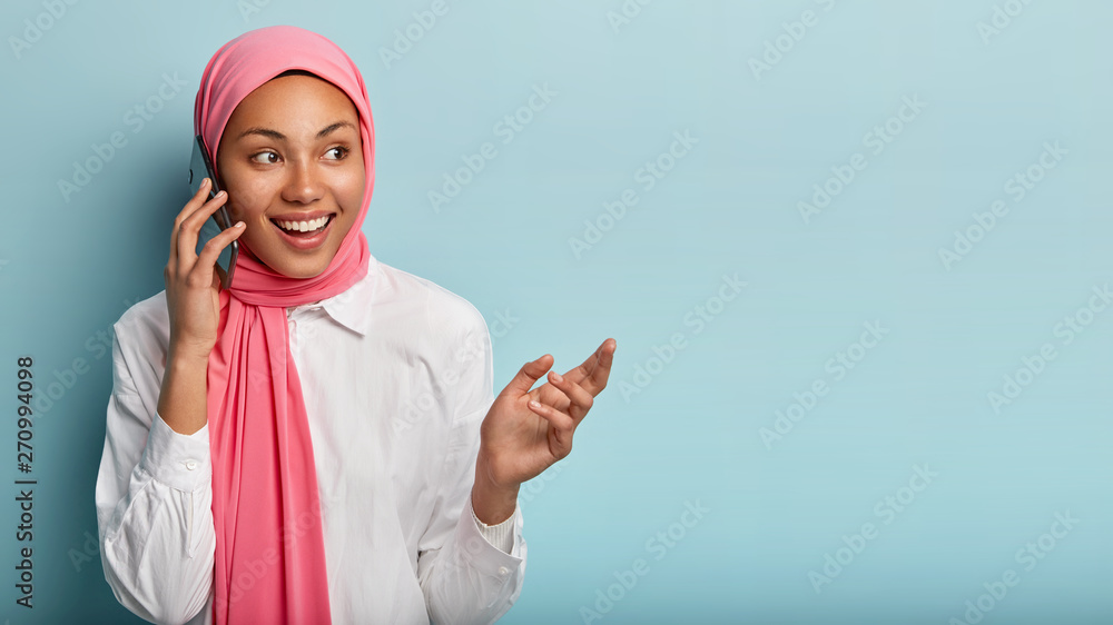 Pretty dark skinned woman talks on mobile phone, enjoys pleasant conversation, wears pink hijab and white shirt, looks aside, isolated over blue backround with blank space. Technology and religion Pretty dark skinned woman talks on mobile phone, enjoys pleasant conversation, wears pink hijab and white shirt, looks aside, isolated over blue backround with blank space. Technology and religion