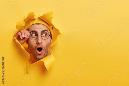 Astonished unshaven man gazes through hole in yellow paper, wears round spectacles, opens mouth with surprisement, copy space to insert text or slogan. Effect of torn paper. Discount and sale