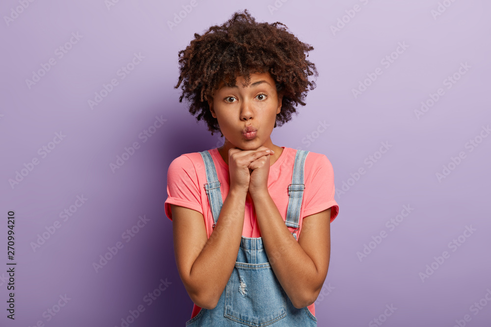 Headshot of lovely woman keeps hands under chin, has folded lips, healthy dark skin, Afro hairstyle, dressed in casual clothes, stands against purple background, wants to kiss beloved person. Headshot of lovely woman keeps hands under chin, has folded lips, healthy dark skin, Afro hairstyle, dressed in casual clothes, stands against purple background, wants to kiss beloved person.
