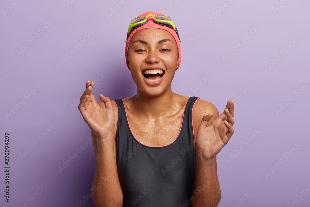 Carefree optimistic dark skinned lady laughs at something funny, clasps hands, being wet after swimming in sea, wears special clothes and equipment, shows white teeth, models indoor. Emotions, rest Carefree optimistic dark skinned lady laughs at something funny, clasps hands, being wet after swimming in sea, wears special clothes and equipment, shows white teeth, models indoor. Emotions, rest