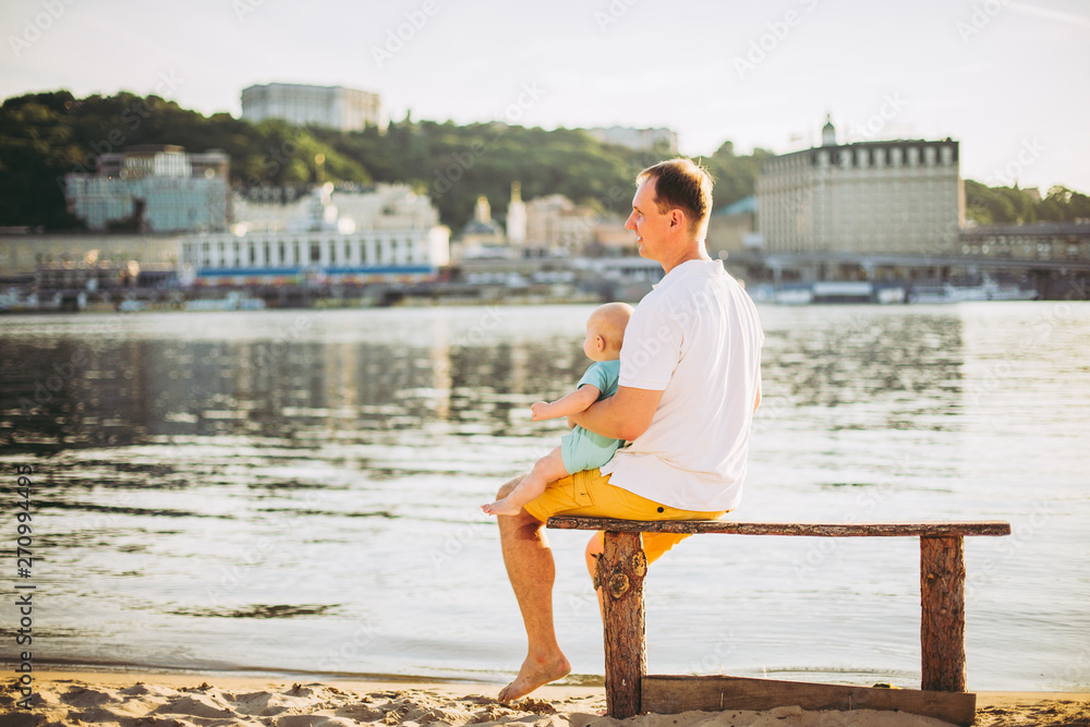 Subject recreation father and little son. Young caucasian dad sits on wooden bench overlooking the town Kiev and holding river of the Dnieper, hugging a toddler baby on sandy beach on the banks pond