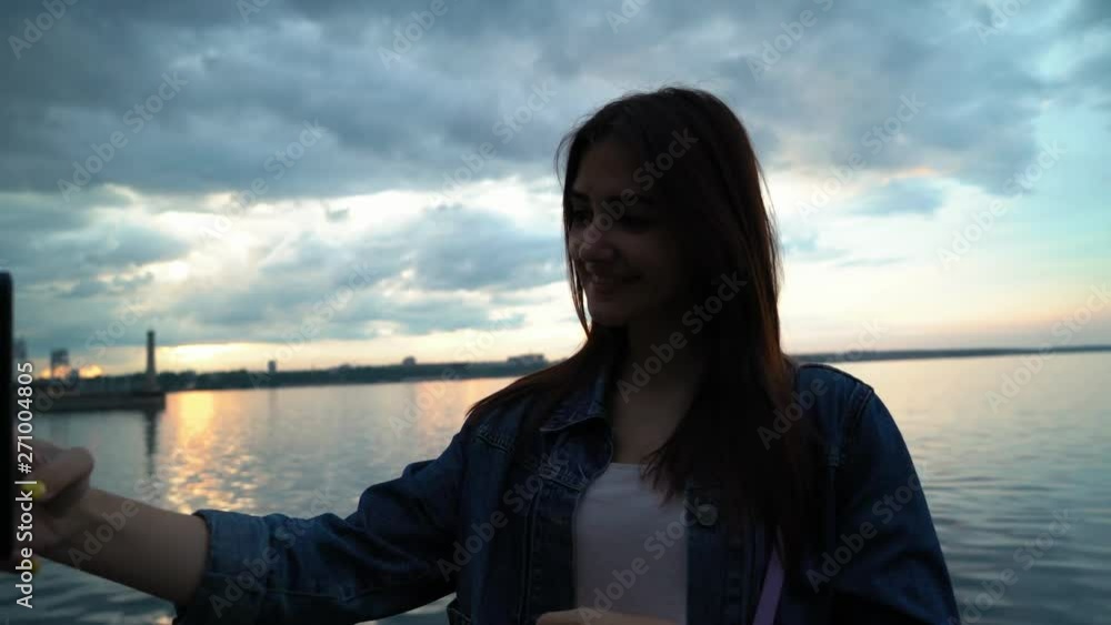 smiling girl doing cool selfies on mobile phone near the water on the quay in the evening at sunset smiling girl doing cool selfies on mobile phone near the water on the quay in the evening at sunset