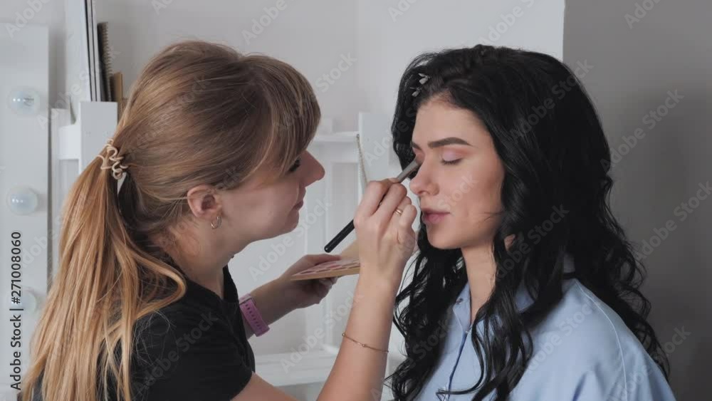 Closeup shot of cheerful young female makeup artist enjoying working with a beautiful young brown haired model, applying fresh trendy makeup on her face with long eyelashes, perfect brows using pencil Closeup shot of cheerful young female makeup artist enjoying working with a beautiful young brown haired model, applying fresh trendy makeup on her face with long eyelashes, perfect brows using pencil