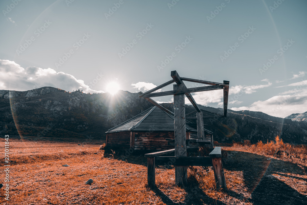 An autumn wide-angle landscape in Altai mountains with a ridge overgrown with fall trees in the background and a glade in the foreground with an abandoned wooden shack and table to eat