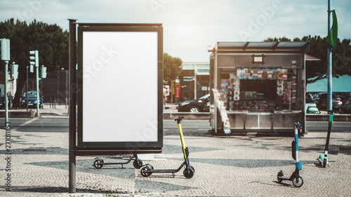 Mockup of an empty urban advert poster surrounded by scooters scattered across the street on the pavement stone; template of a blank street information billboard with a newsstand in the background photo