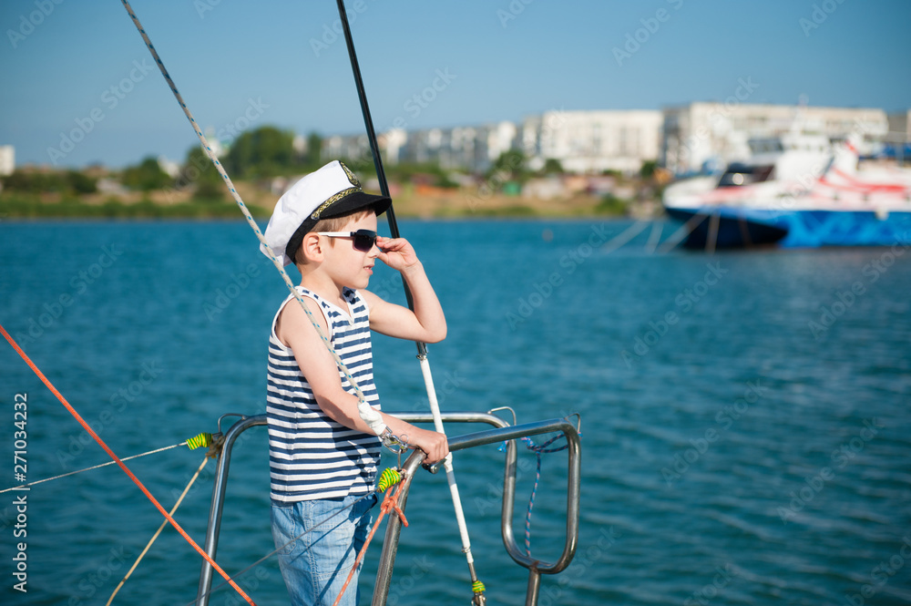 happy caucasian little boy in white captain hat and striped tank top with denim shorts standing on luxury yacht board adjusting sunglasses in sea port during summer travel