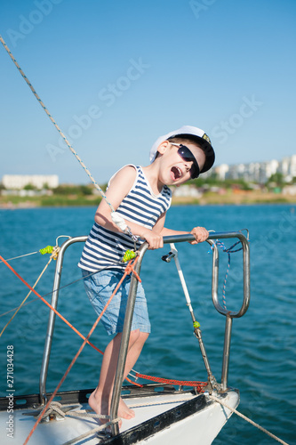 funny delightful little boy wearing sunglasses and captain hat fooling around on ocean yacht board during summer travel vacation cruise in sea port