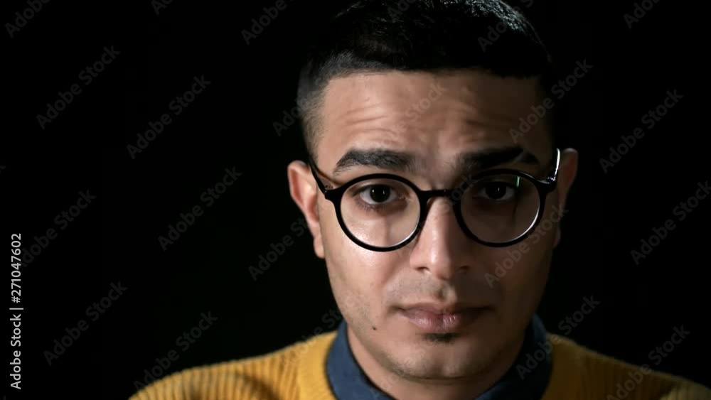 Close up of young Arab man putting on round shape glasses and looking at camera with serious expression on his face Close up of young Arab man putting on round shape glasses and looking at camera with serious expression on his face